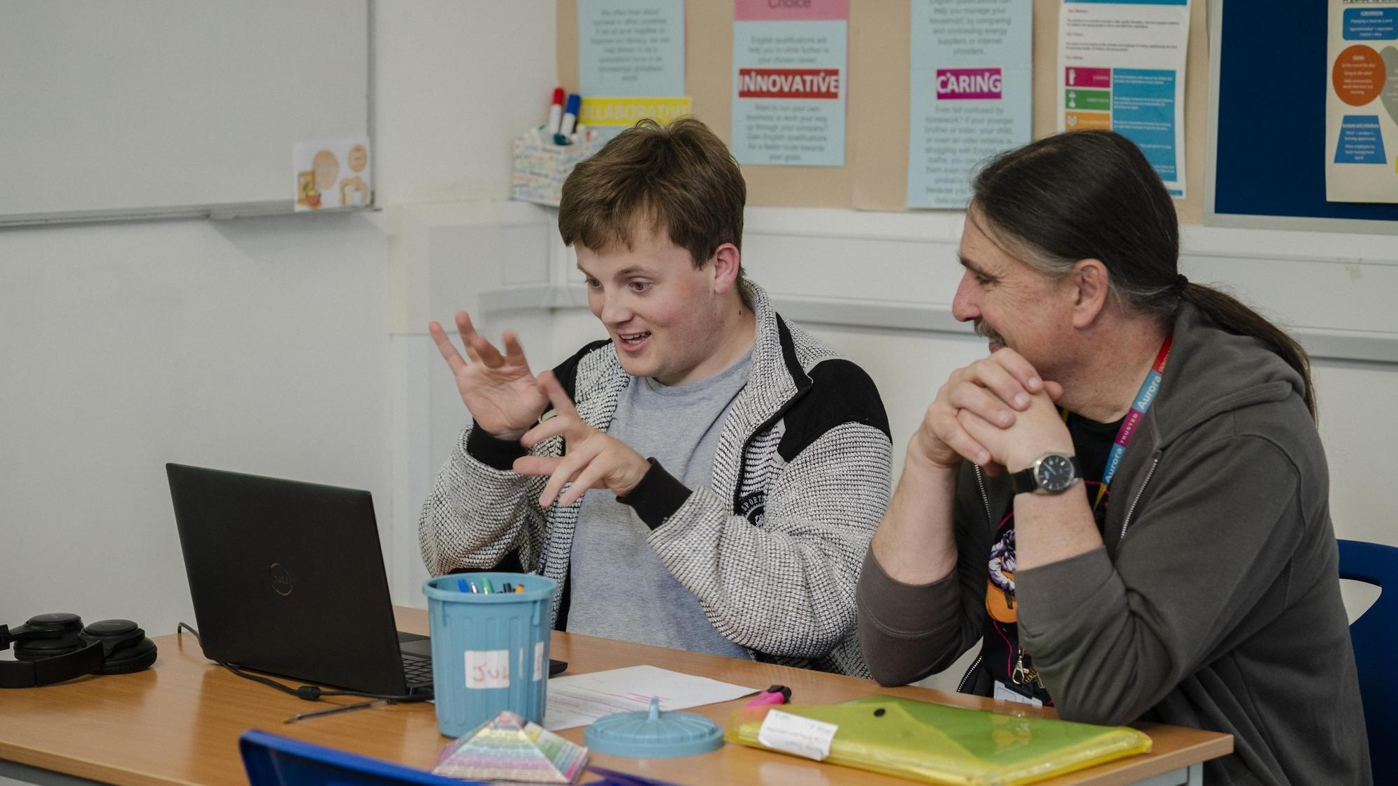 Student and their teacher in a classroom looking at a laptop together with pens, pencils and paper on the desk