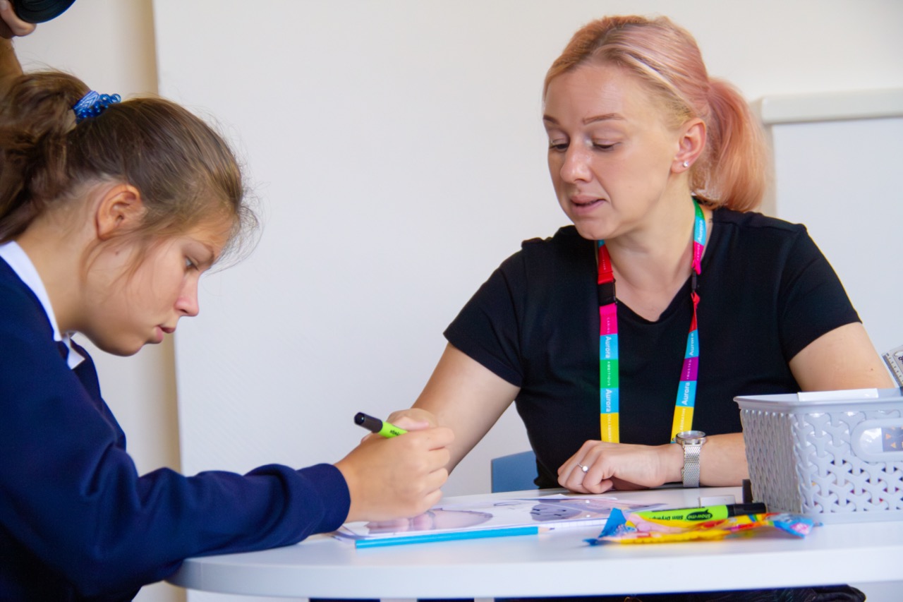 Student and their teacher in a classroom looking at a laptop together with pens, pencils and paper on the desk