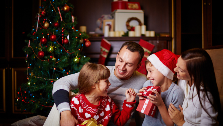 family of four sitting on the floor next to their christmas tree, opening presents and smiling
